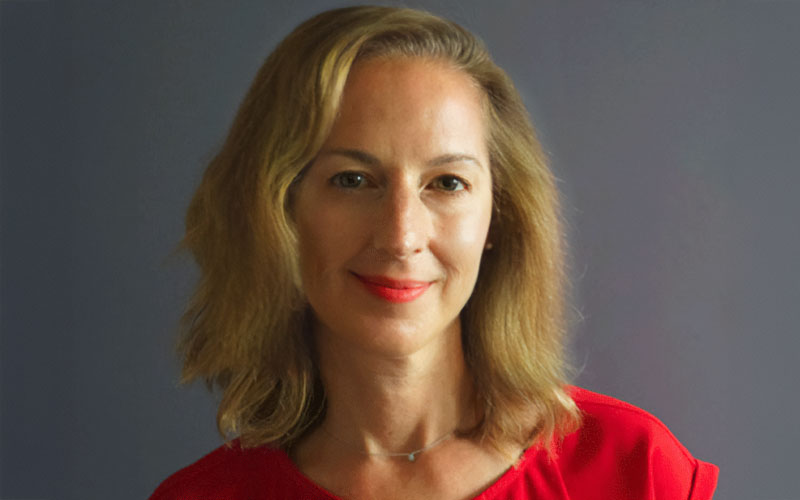 Professional headshot of a smiling woman with shoulder-length blonde hair, wearing a red top, against a neutral background.
